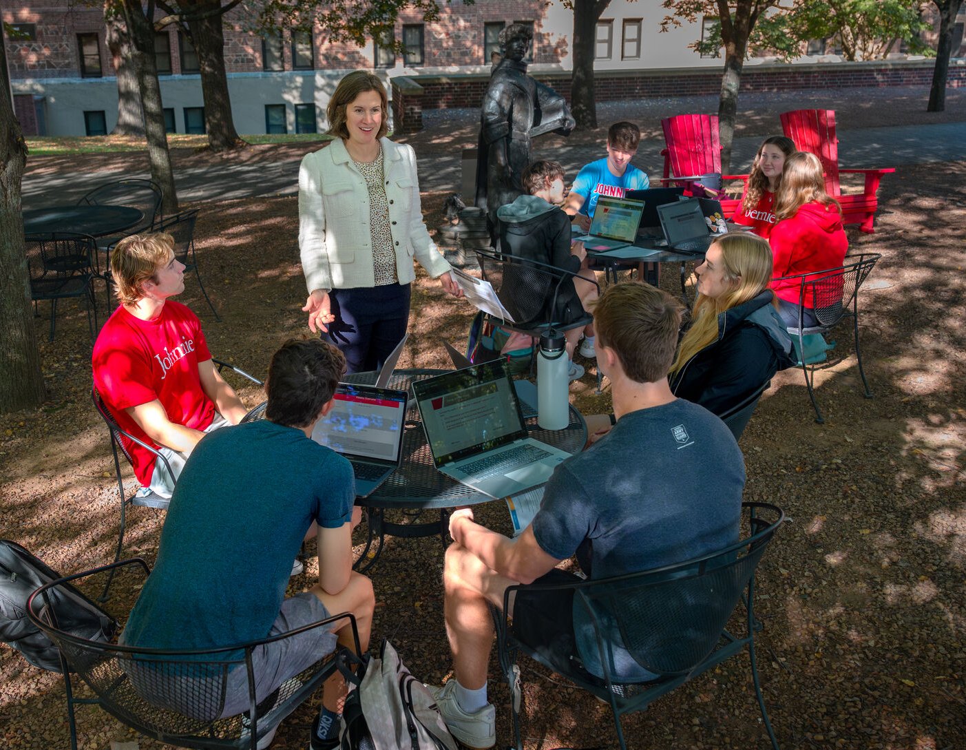 A group of college students sit outdoors at round tables with laptops, engaging in discussion with a standing woman, likely a professor, under the shade of trees on a sunny day.