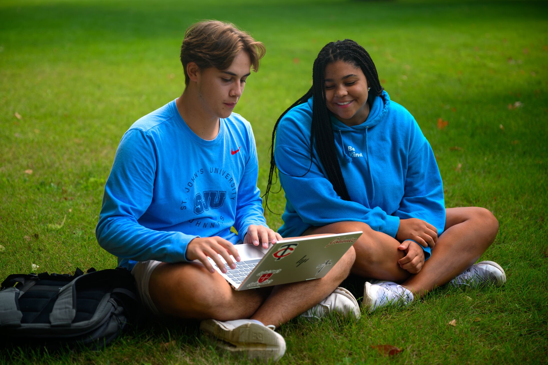 Two students sit on grass with a laptop, both wearing blue sweatshirts. One types on the laptop while the other looks on and smiles. A black backpack lies nearby. The scene is bright and outdoors.