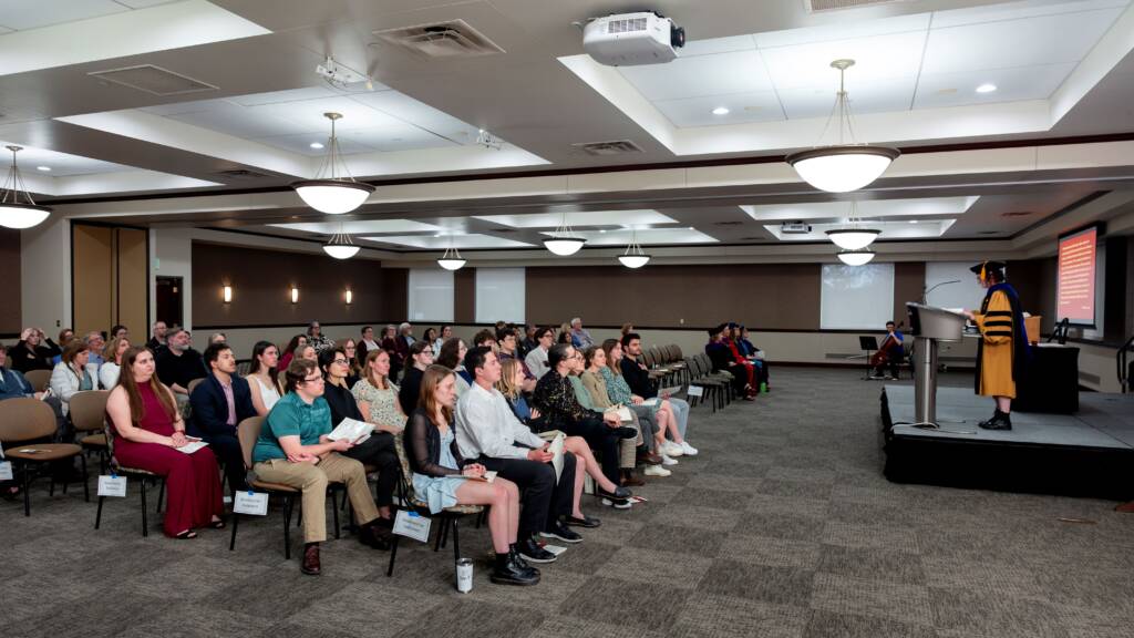 A speaker in academic regalia addresses an audience seated in rows inside a large, well-lit conference room. People attentively watch the speaker, and a presentation is projected onto a screen at the front.