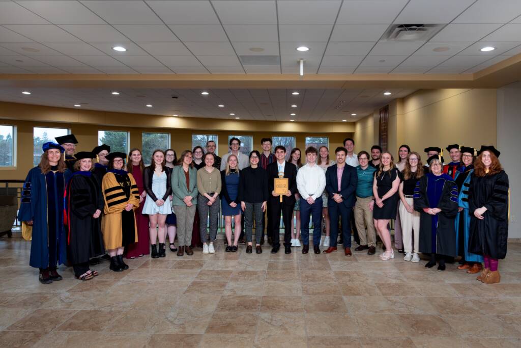 A large group of students and faculty, some in academic robes, pose together indoors on a tiled floor for a graduation or award ceremony. One person in the center holds a wooden plaque.