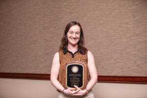 A woman with brown hair, wearing a sleeveless, patterned blouse, stands indoors holding a black and gold award plaque, smiling at the camera. A tan and brown textured wall is in the background.