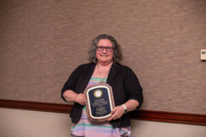 A person with curly gray hair and glasses, wearing a striped dress and black jacket, smiles while holding a plaque award against a textured beige and brown wall.