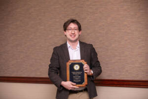 A man wearing glasses and a brown blazer stands indoors, smiling and holding a wooden plaque award in front of a beige textured wall.