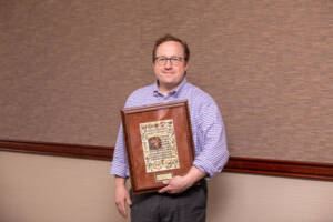 A man wearing glasses and a checkered shirt stands indoors, smiling and holding a large framed award with decorative artwork and a small gold name plaque.