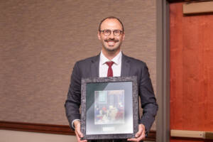 A man in a suit and red tie stands indoors, smiling and holding a framed artwork with both hands. The background features a beige patterned wall and a wooden door.