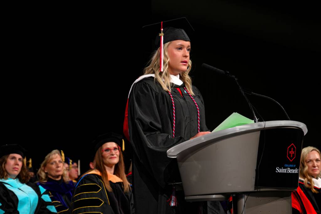 A student in a graduation cap and gown speaks at a podium during a commencement ceremony, with faculty members seated in academic regalia behind her.