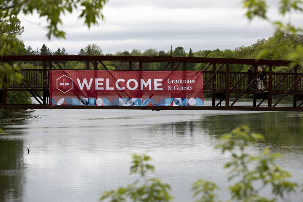 A large red banner reading “Welcome Graduates & Guests” hangs on a pedestrian bridge over a calm river, surrounded by green trees and foliage.