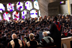 A person in academic regalia speaks at a podium to a large audience of graduates and faculty during a ceremony in a hall with colorful stained glass windows.