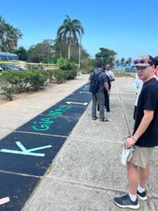 A young man in sunglasses stands on a sidewalk near large chalk letters while several people walk ahead. Palm trees and clear blue sky are in the background, creating a tropical setting.