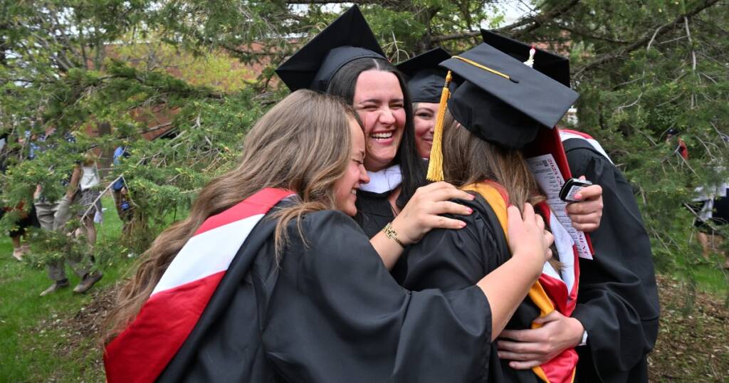 Four graduates in caps and gowns embrace and smile joyfully outdoors, surrounded by green trees, celebrating together.