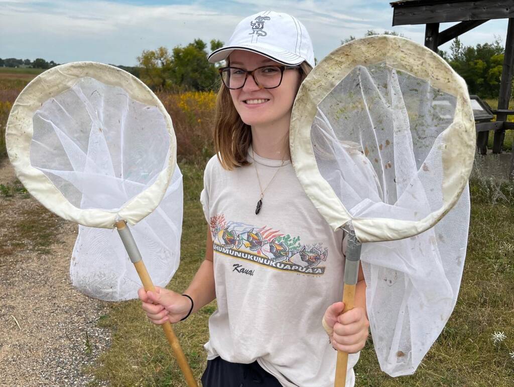 A smiling person wearing glasses, a white cap, and a T-shirt stands outdoors holding two large insect nets, with grassy fields and yellow wildflowers in the background.