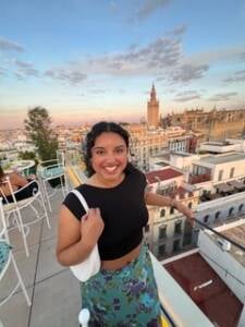 A woman with dark curly hair, a black crop top, and a floral skirt smiles on a rooftop terrace overlooking a cityscape with historic buildings at sunset.