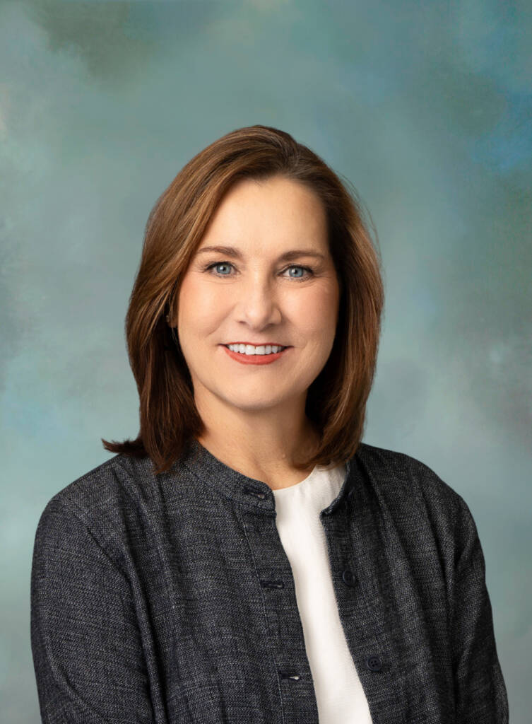 A woman with straight, shoulder-length brown hair and blue eyes smiles at the camera. She wears a dark grey textured jacket over a white top, posed against a soft blue and grey background.