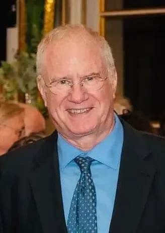 An older man with short white hair and glasses smiles at the camera. He is wearing a blue dress shirt, blue patterned tie, and a dark suit jacket. The background is blurred with other people and some greenery visible.