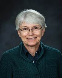 An older woman with short gray hair and glasses smiles at the camera. She is wearing a dark green sweater over a dark collared shirt, with a dark background behind her.