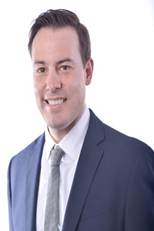 A man wearing a navy blue suit, white shirt, and light gray tie smiles at the camera against a plain white background.