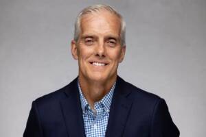 A middle-aged man with short gray hair wearing a navy blazer and a blue-and-white checkered shirt smiles at the camera against a plain gray background.