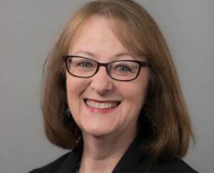 A woman with shoulder-length light brown hair and glasses smiles at the camera. She is wearing a black top and the background is plain and light gray.