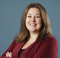 A woman with long brown hair, wearing a maroon cardigan over a black top, stands smiling with arms crossed in front of a plain blue background.