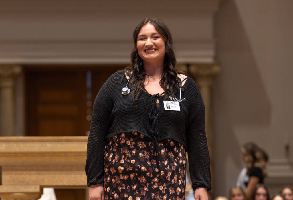 A woman with long brown hair, wearing a black top, floral skirt, stethoscope, and ID badge, smiles while standing indoors in a well-lit room with columns in the background.