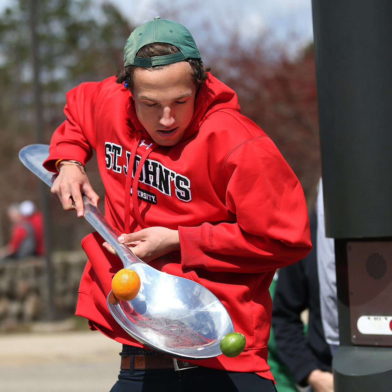 A person in a red St. John's University hoodie and green backwards cap balances two limes and an orange on a giant silver spoon outdoors.