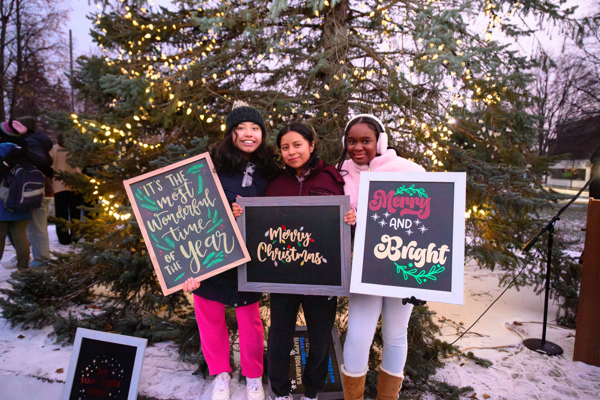 Three girls in winter clothes stand smiling in front of a lit Christmas tree, each holding colorful holiday-themed signs that read “It’s the most wonderful time of the year,” “Merry Christmas,” and “Merry and Bright.”.