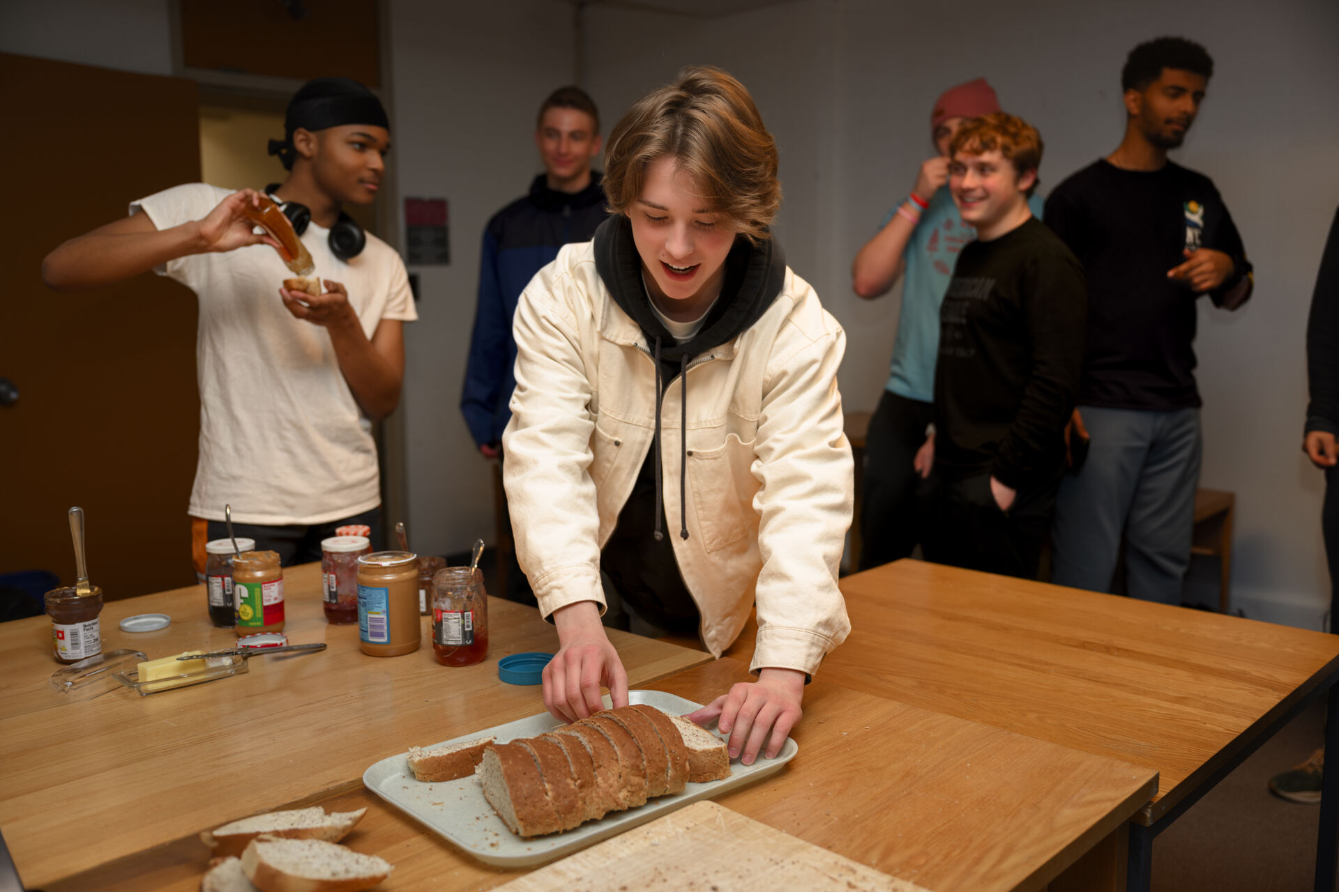 A group of young people stand around a table. One person in a white jacket slices bread, while another spreads something on a slice. Jars of spreads and utensils are on the table. Everyone looks happy and engaged.