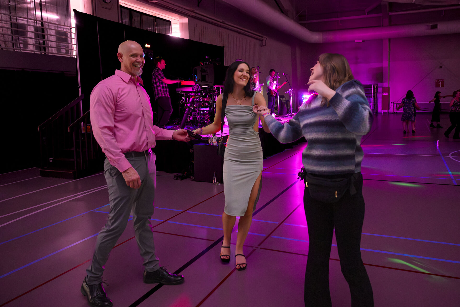 Three people are dancing and smiling together in a gymnasium with purple lighting. A live band plays on stage in the background, and a few other people are visible dancing in the distance.