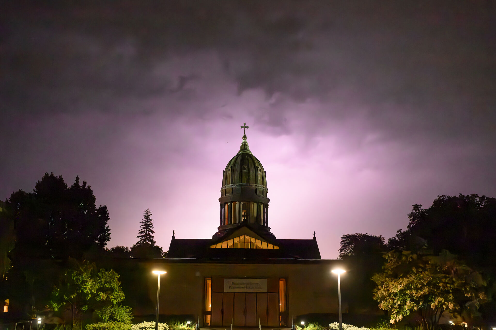 A church with a large dome and cross on top is illuminated against a dramatic, cloudy night sky. Trees and glowing streetlights surround the building, creating a silhouette effect.