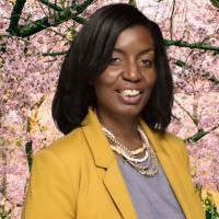 Kristen Hodge-Clark, a woman with shoulder-length dark hair, wearing a yellow blazer, gray top, and layered necklace, smiling in front of pink blossoming trees.
