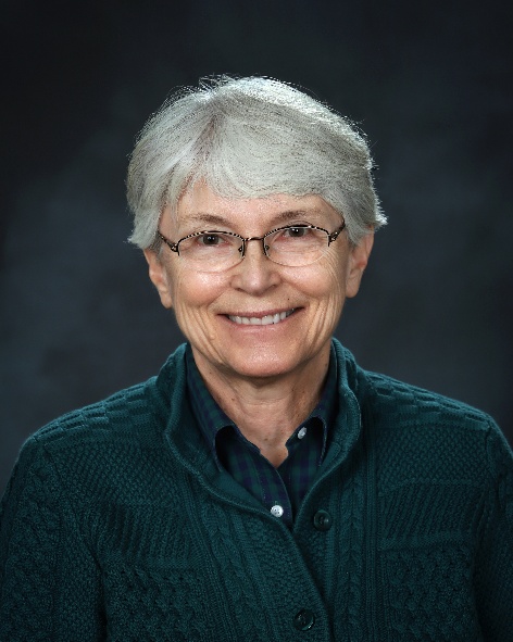 Jeanne Marie Lust ’73, OSB, an older woman with short gray hair and glasses, smiling at the camera. She is wearing a dark green textured sweater over a collared shirt, with a dark background behind her.