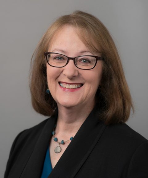 Pam Wheelock, a middle-aged woman with shoulder-length brown hair and glasses, smiling, wearing a black blazer, teal top, and a beaded necklace, posed against a plain gray background.