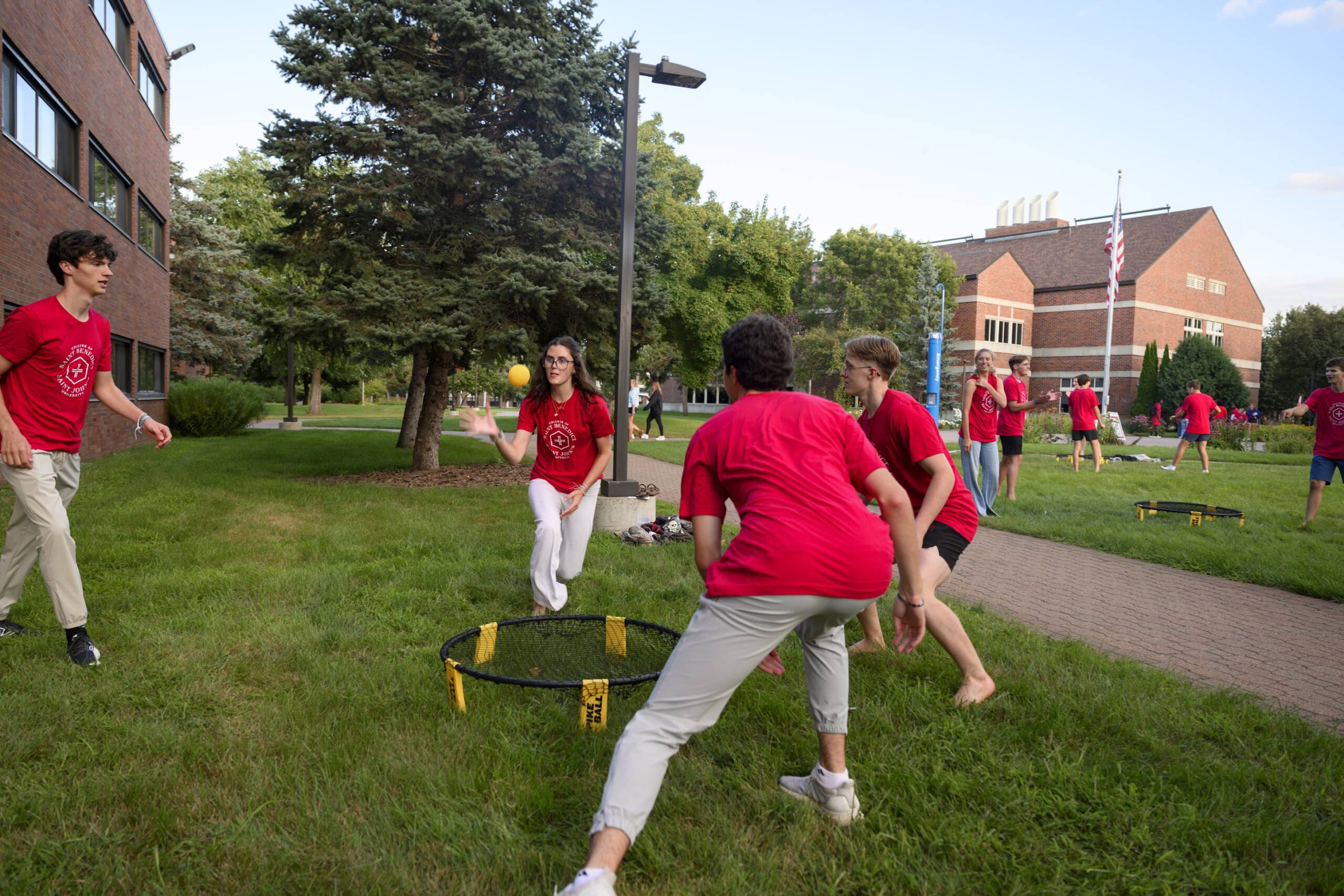 A group of young people in red shirts play Spikeball on a grassy area between brick buildings, with others in the background and trees surrounding the scene on a sunny day.