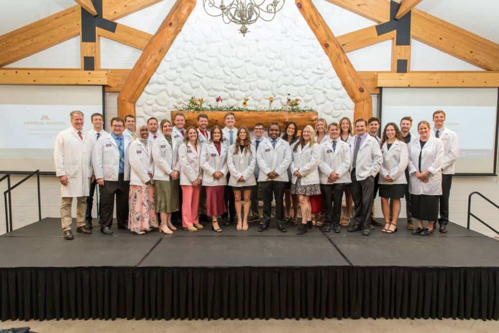 A group of people in white lab coats stand together on a stage, posing for a group photo in a large room with wooden beams and a white textured wall behind them.