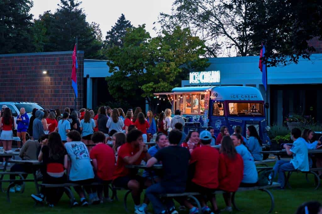 A large group of people gather outdoors in the evening, some sitting at picnic tables and others forming a line at a brightly lit ice cream truck parked on the grass. Trees and a building are in the background.