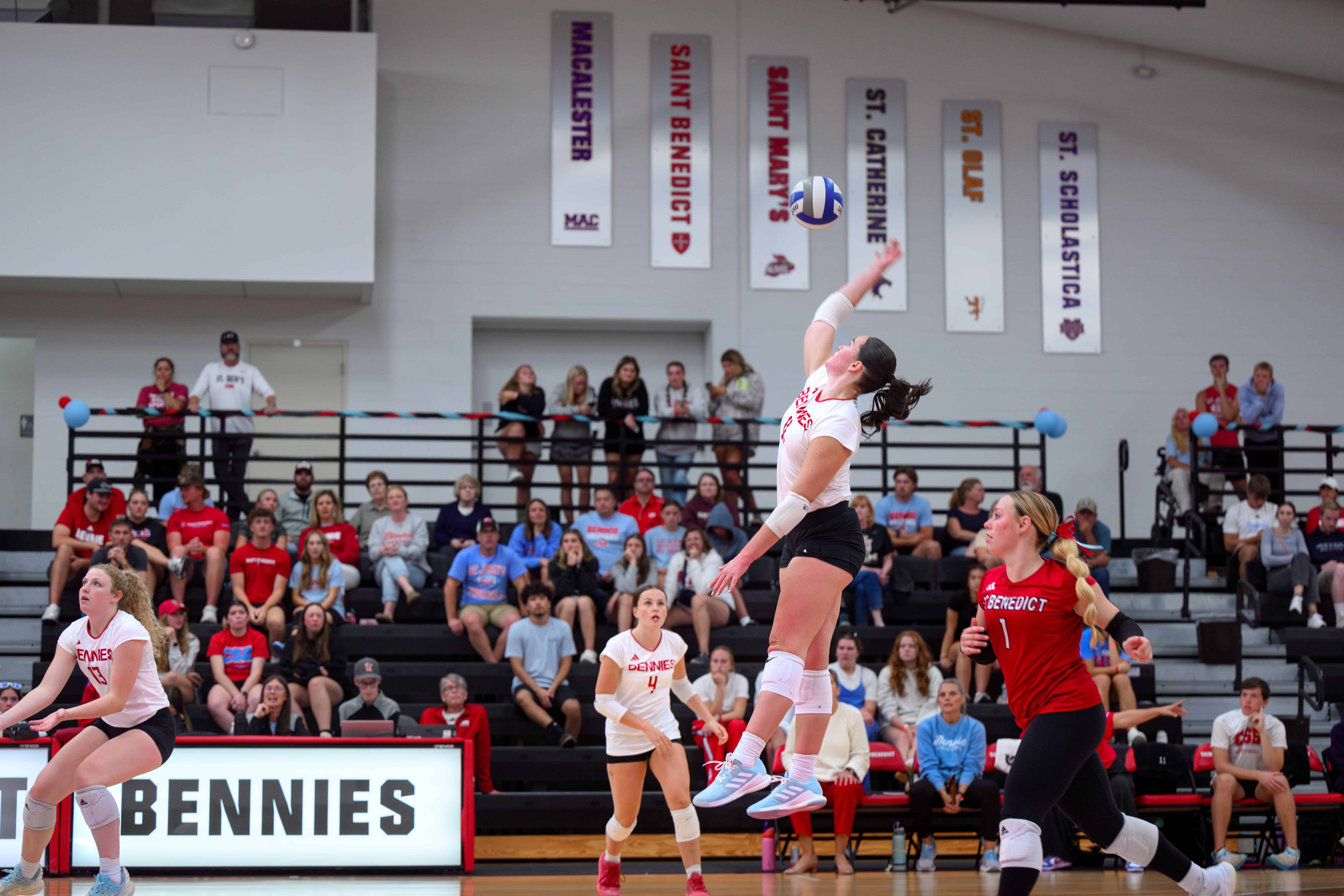 A volleyball player in a white jersey jumps to spike the ball while teammates and an opponent watch. Spectators sit in bleachers behind the court, and banners hang on the wall above them.