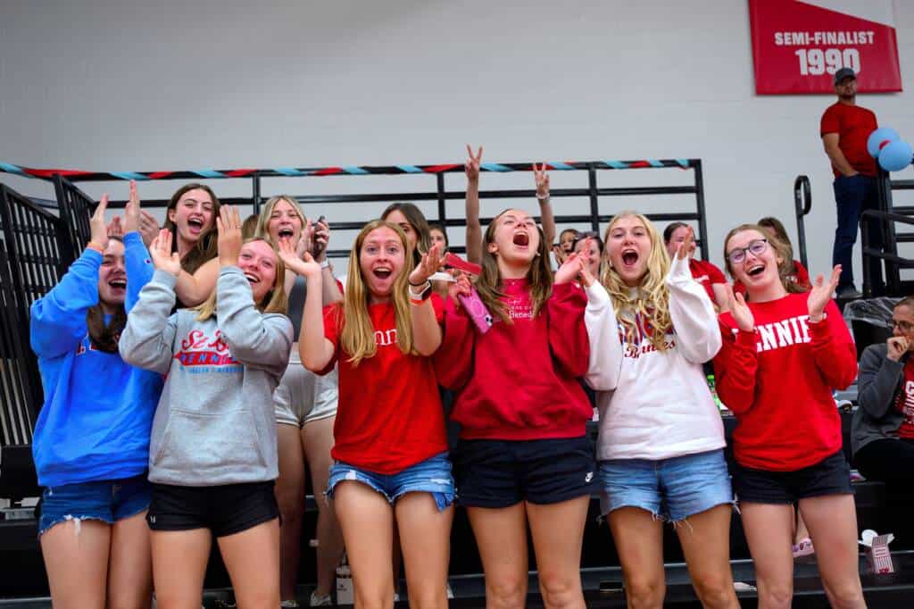 A group of enthusiastic teenage girls in casual shorts and sweatshirts cheer and clap in bleachers at a school event, with a red "Semi-Finalist 1990" banner hanging in the background.