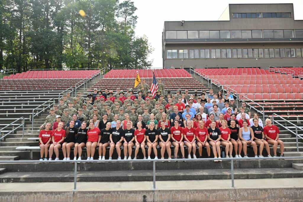 A large group photo on stadium bleachers shows people in military uniforms standing in rows behind individuals in red, black, and white athletic shirts sitting in front, with trees and a building in the background.