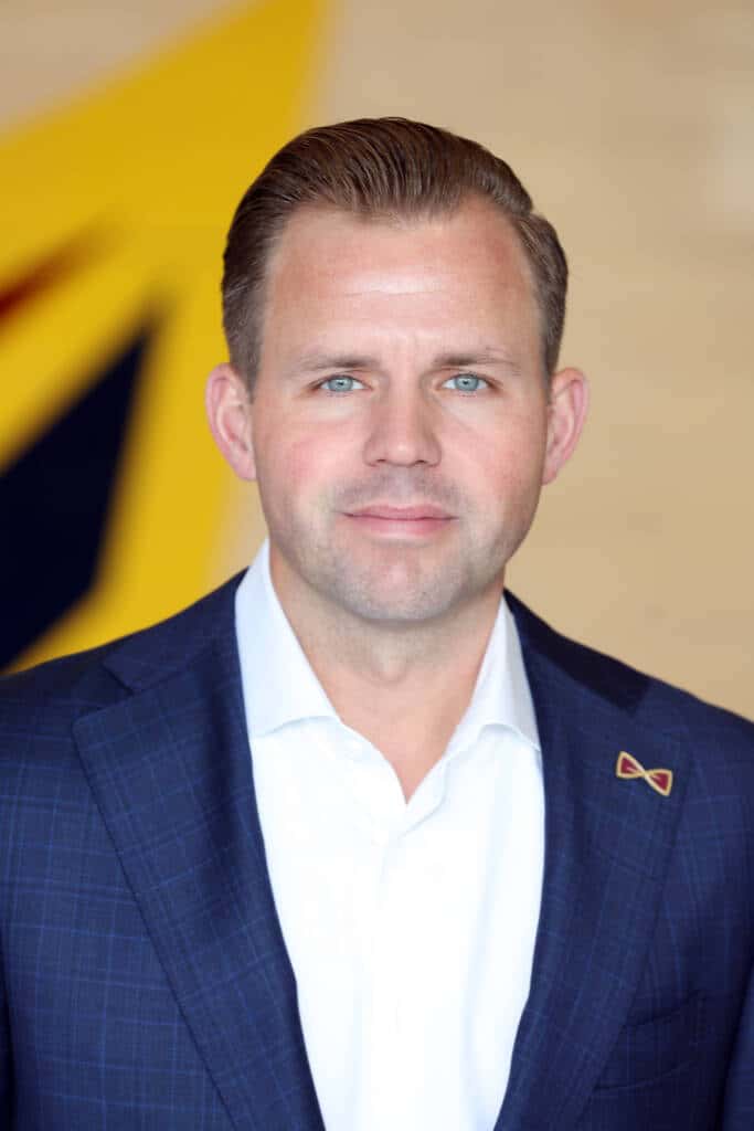 A man wearing a blue checked suit jacket and white shirt stands in front of a blurred yellow and dark blue background, looking at the camera with a neutral expression.