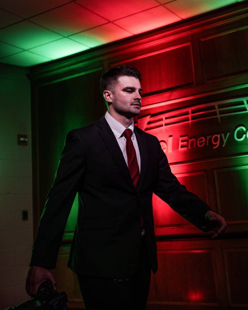 A man in a black suit with a red tie stands indoors against a wooden wall, lit by dramatic red and green lighting. He holds something in his left hand and looks forward with a serious expression.