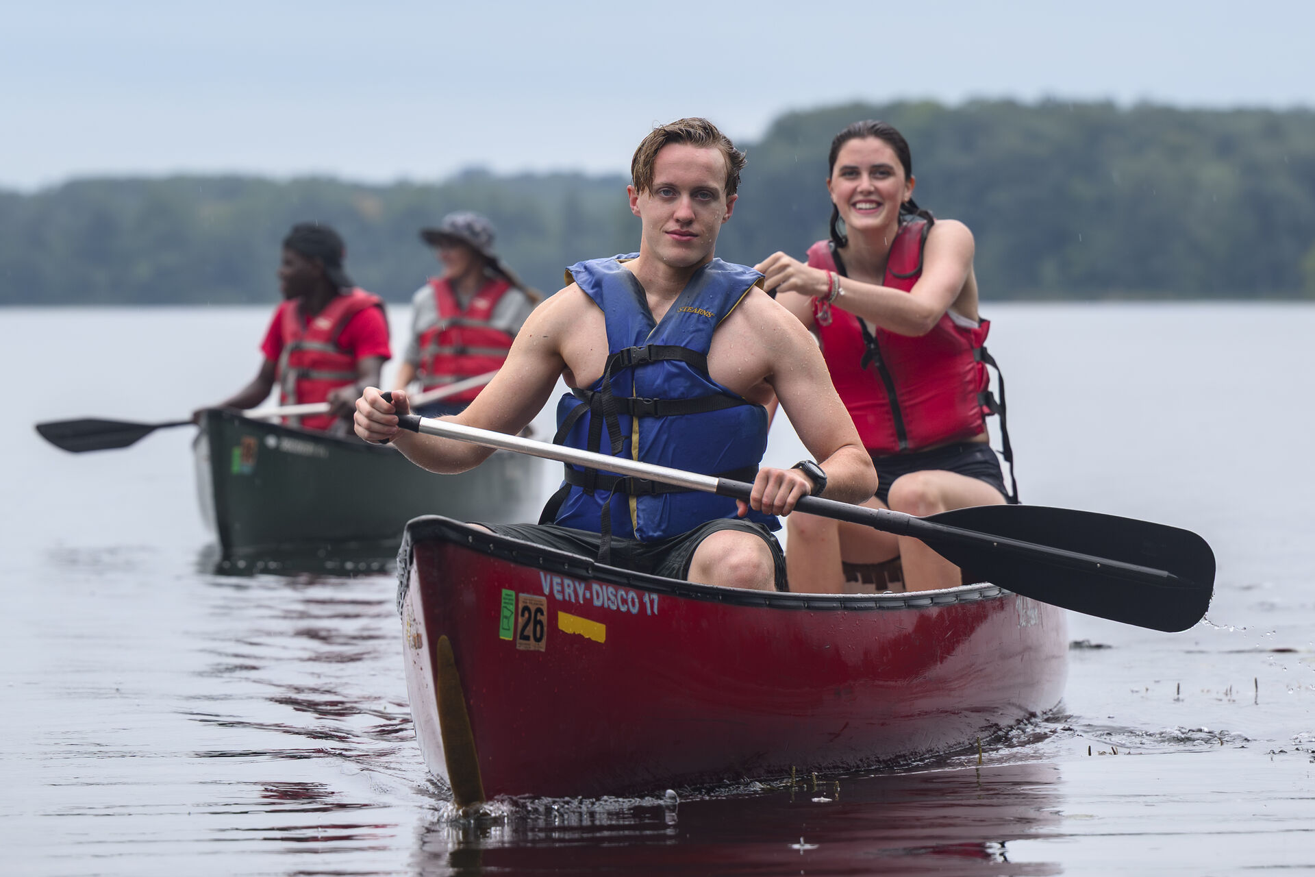 Two people paddle a red canoe on a calm lake, both wearing life jackets. Another canoe with two people is visible in the background. Trees and a cloudy sky are in the distance.