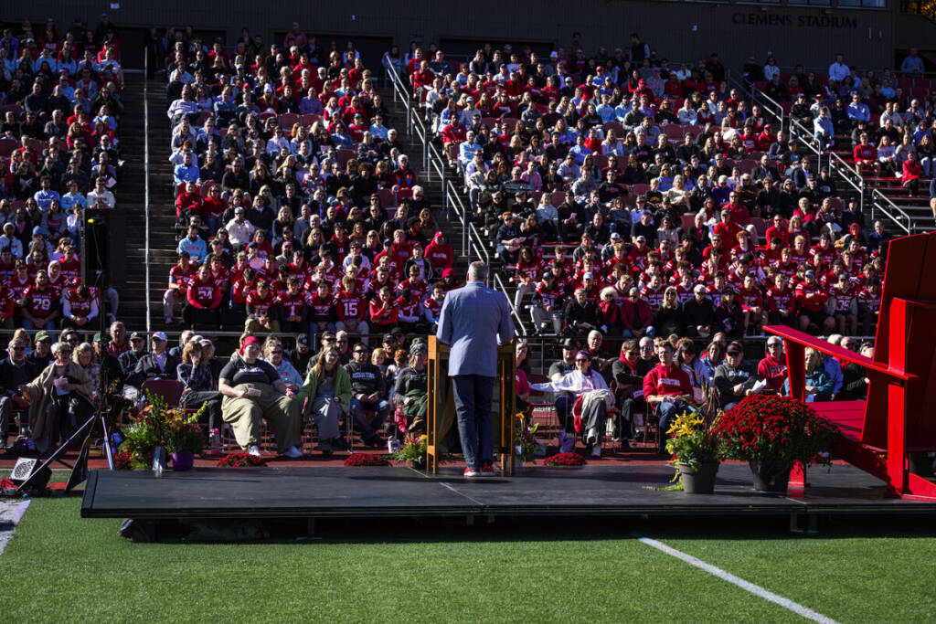 A person stands at a podium on a stage addressing a large crowd seated in stadium stands; most people are wearing red, and there are flowers decorating the stage.