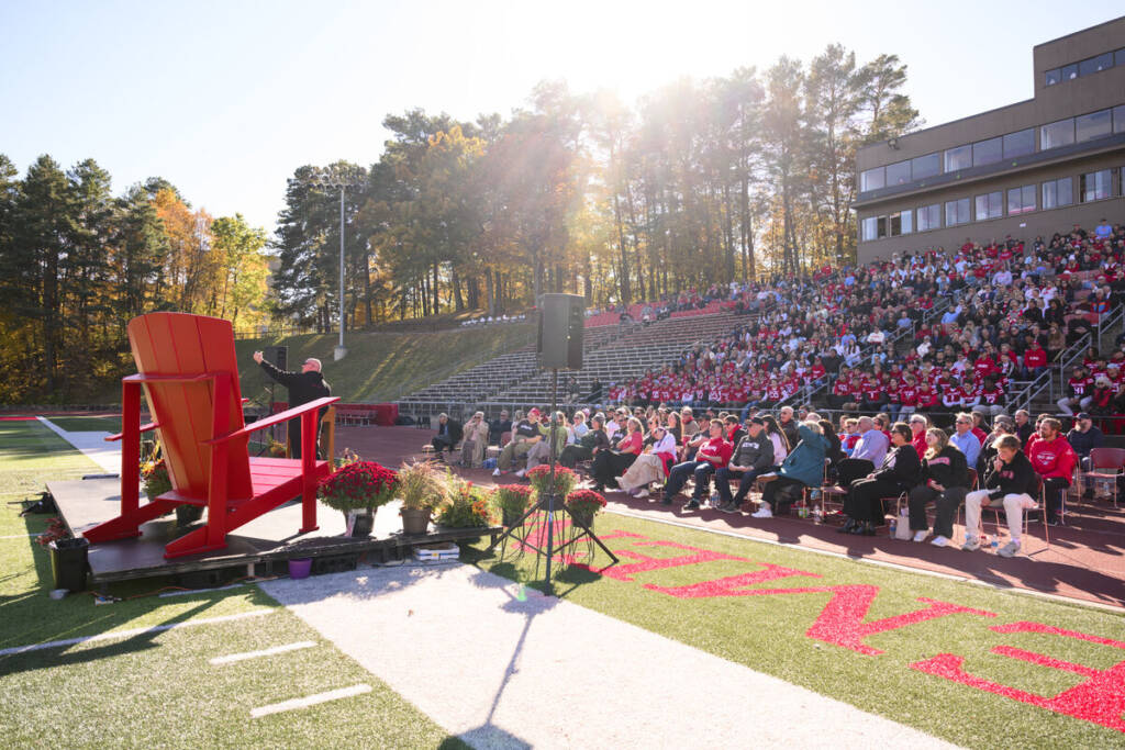 A speaker addresses a large crowd seated in stadium bleachers at an outdoor event on a sunny day; autumn trees and a modern building are visible in the background.