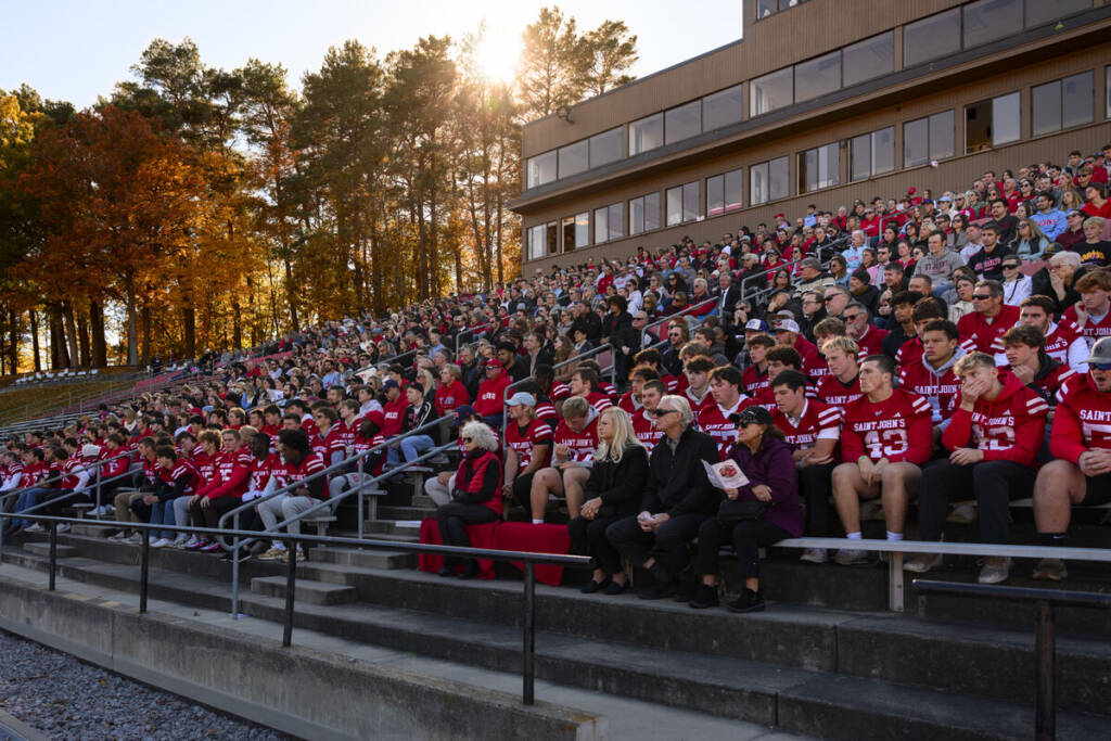 A large crowd sits in outdoor stadium bleachers, many wearing red sports jerseys, with autumn trees and sunlight in the background. Some people hold programs, and most are focused on an event out of view.