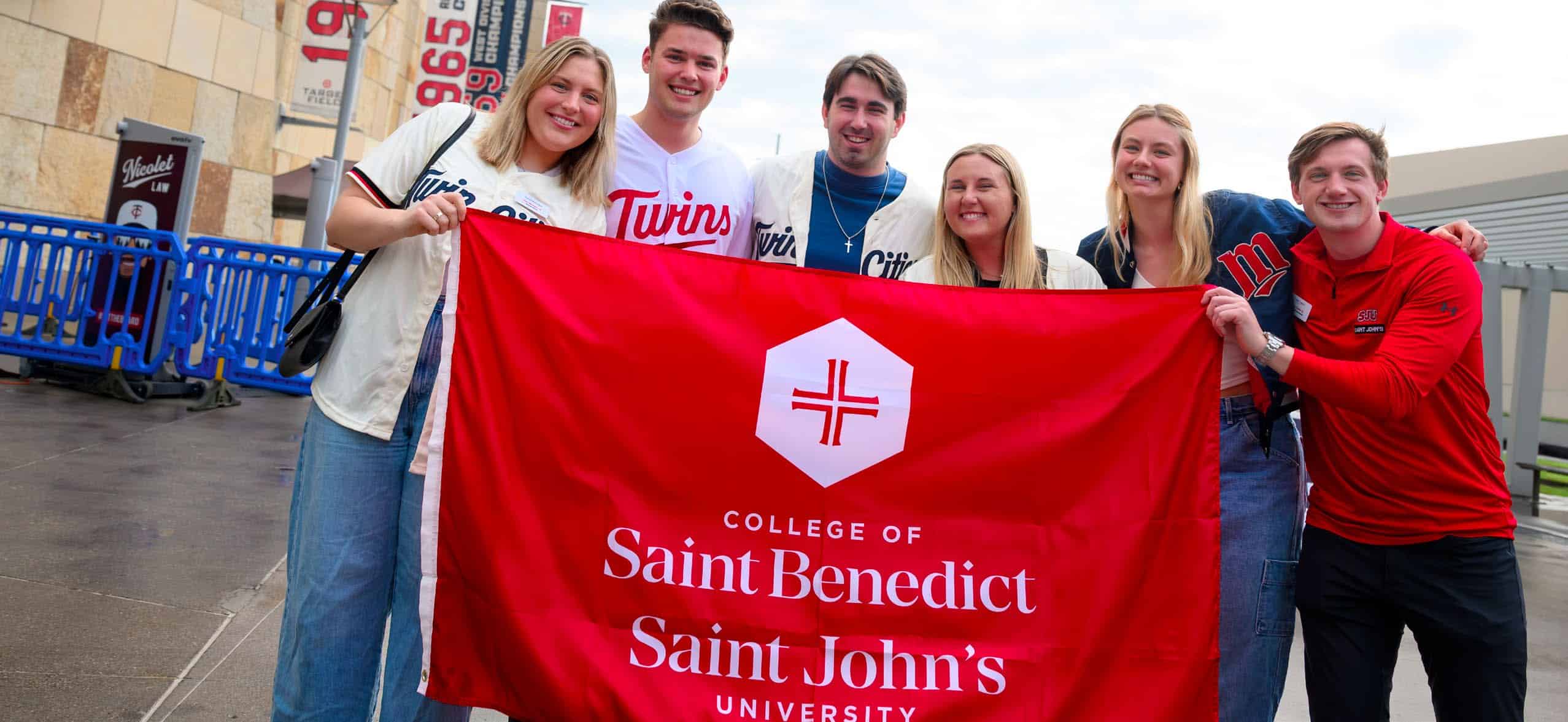 Six smiling young adults stand outdoors holding a large red banner that reads “College of Saint Benedict Saint John’s University.” Some are wearing sports jerseys. They are posing together in a casual, cheerful group.