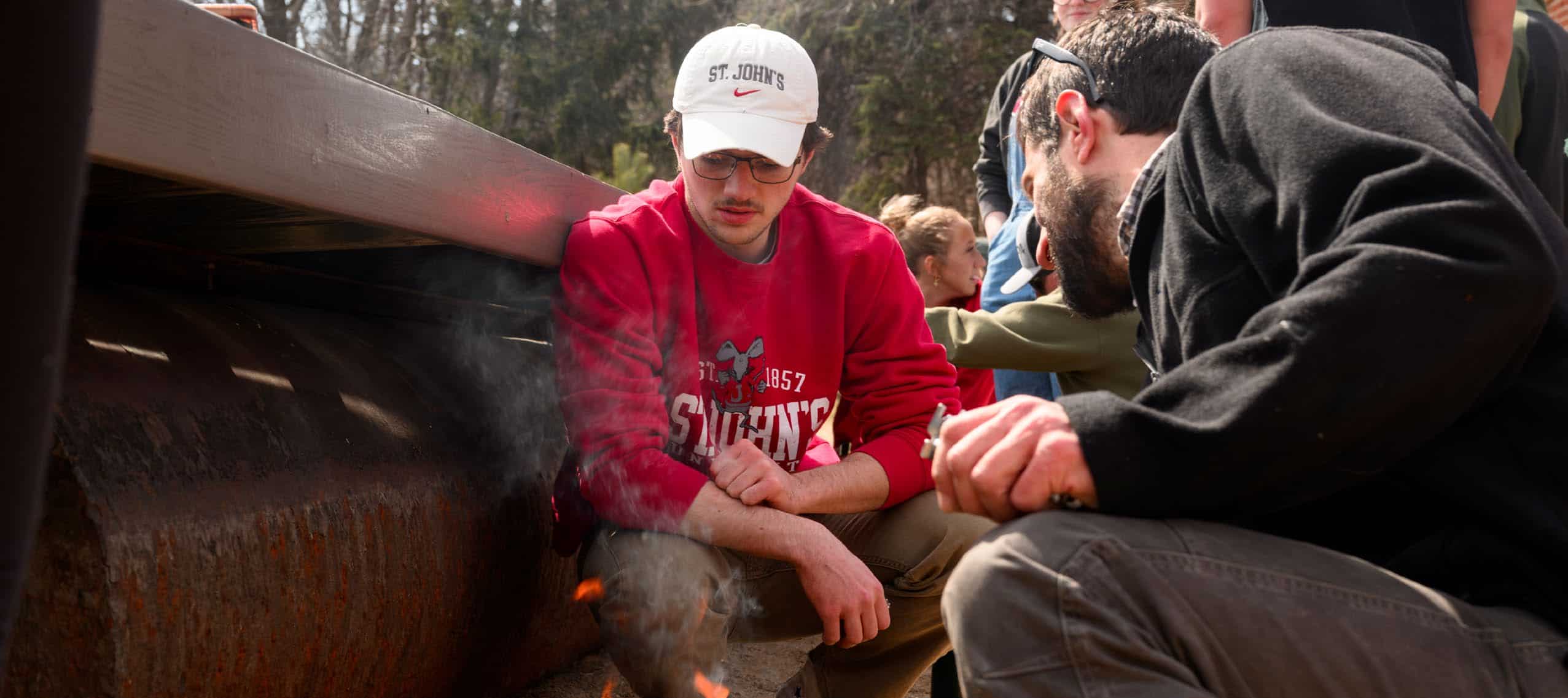 Two people crouch near a small fire outdoors, one wearing a red sweatshirt and white cap. Other people stand nearby, watching the activity attentively. Smoke rises between the crouching individuals.