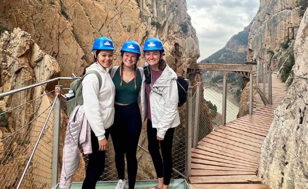 Three women wearing blue helmets and outdoor clothing stand smiling on a glass platform along a canyon walkway, with rocky cliffs and a winding river visible in the background.