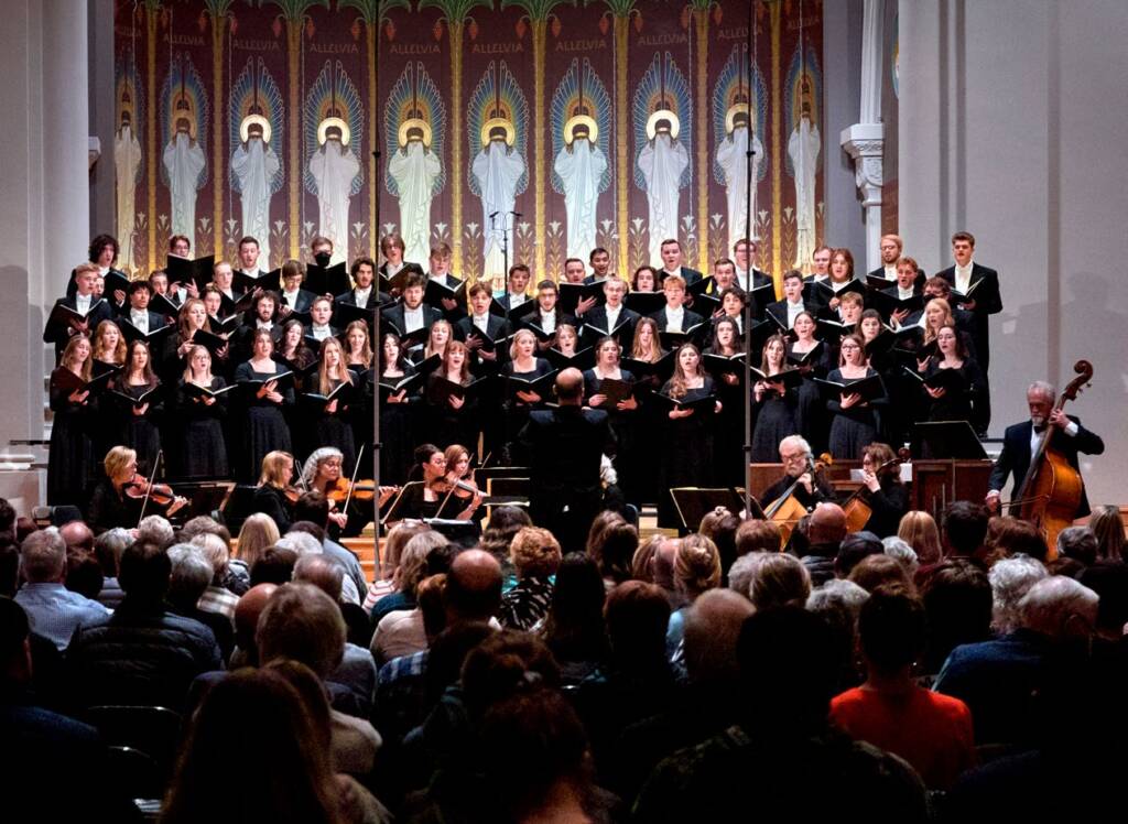 A large choir and orchestra perform on stage in a church or concert hall, facing an audience. The ornate backdrop features tall arches and angel murals. The conductor stands in front, leading the musicians and singers.