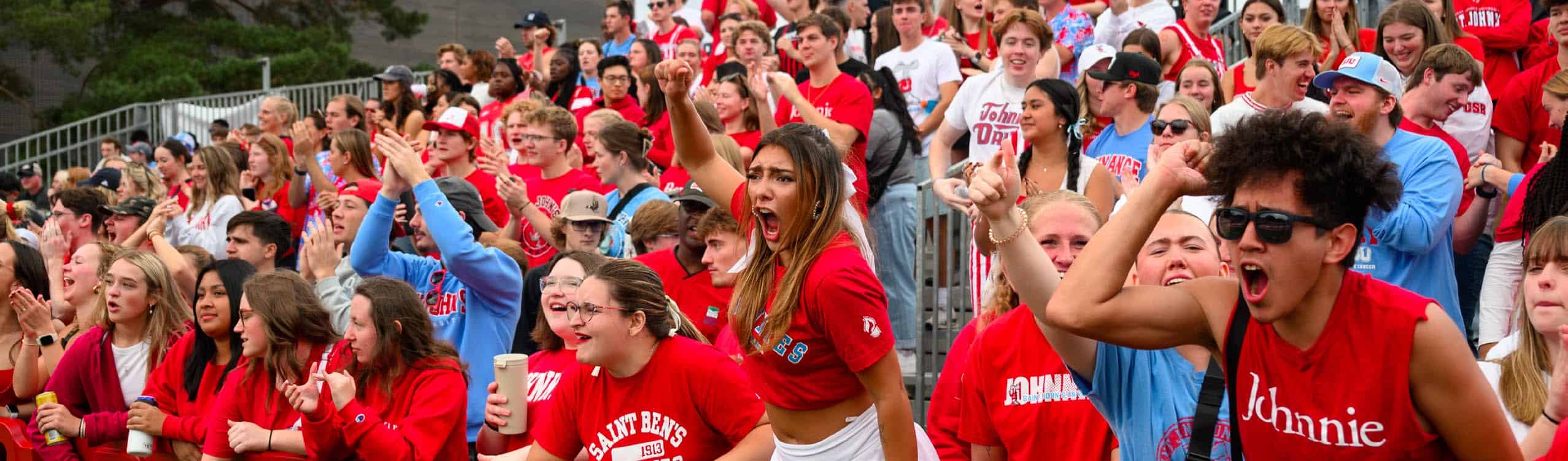 A large, energetic crowd of young people dressed in red and blue shirts cheer passionately at an outdoor event, showing excitement and enthusiasm.