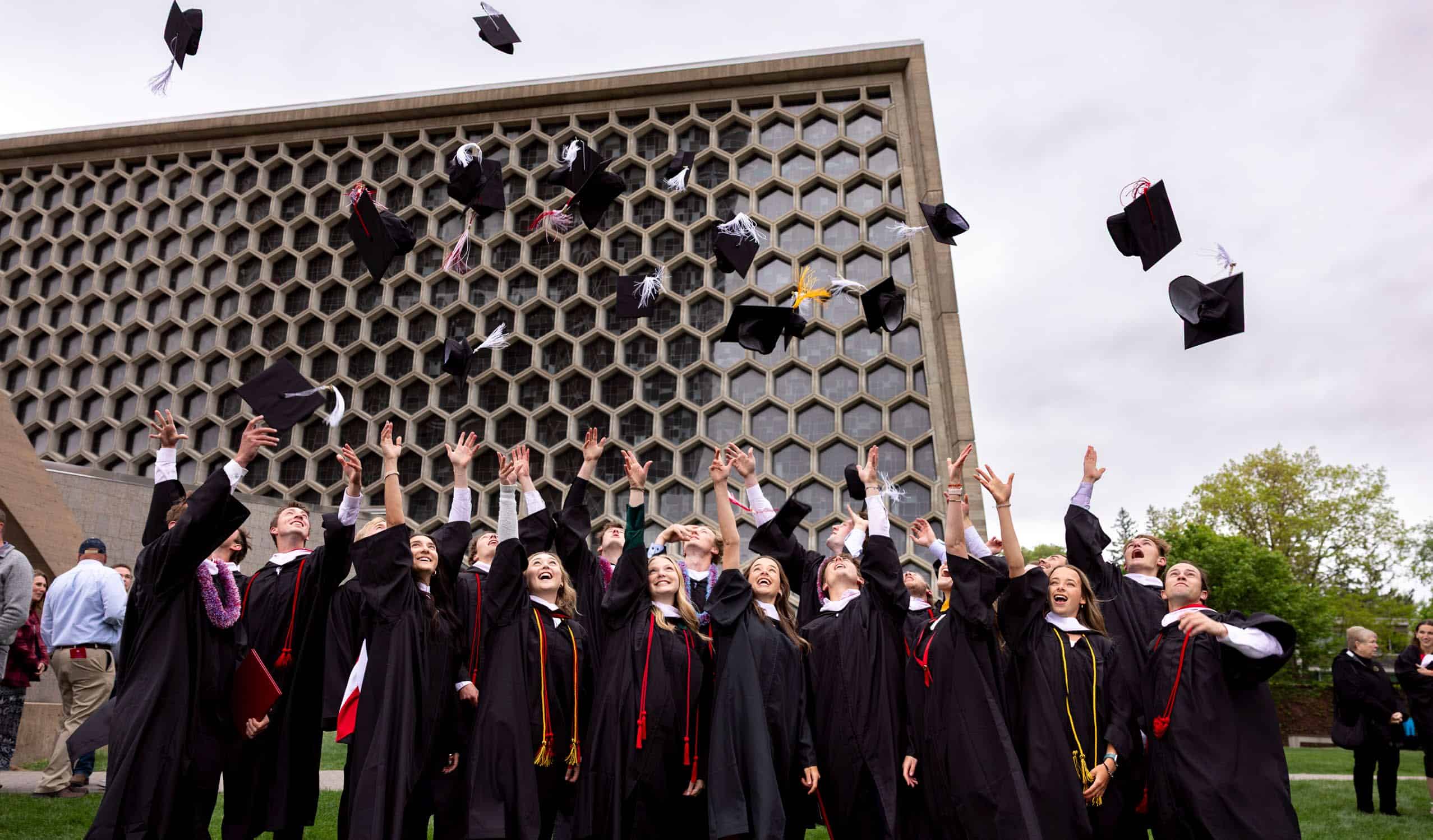 A group of graduates in caps and gowns celebrate by tossing their caps into the air outside a modern building with a honeycomb-patterned facade on a cloudy day.